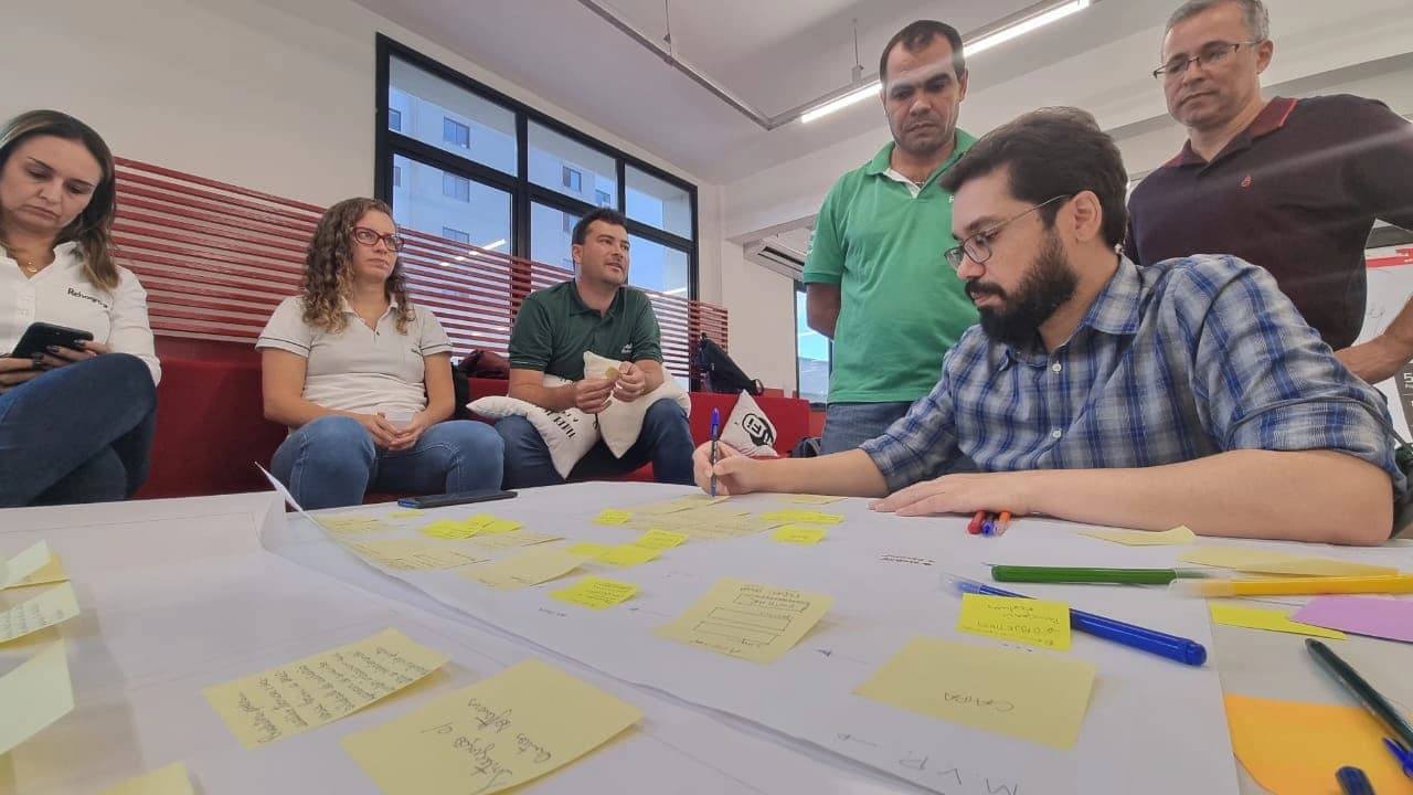 Participante escrevendo em post-it durante dinâmica do Design Sprint, com colegas ao redor da mesa.