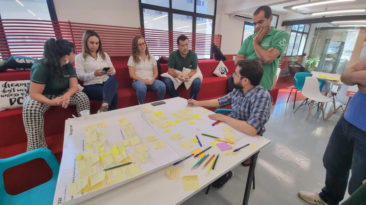 Equipe reunida ao redor de painel com post-its durante o Design Sprint, em espaço de trabalho aberto.