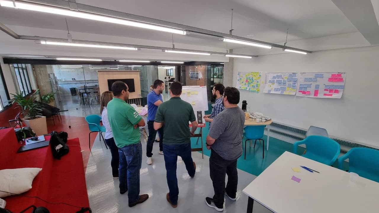 Participantes em pé debatendo em frente a um quadro com post-its, com painéis de atividades na parede ao fundo.