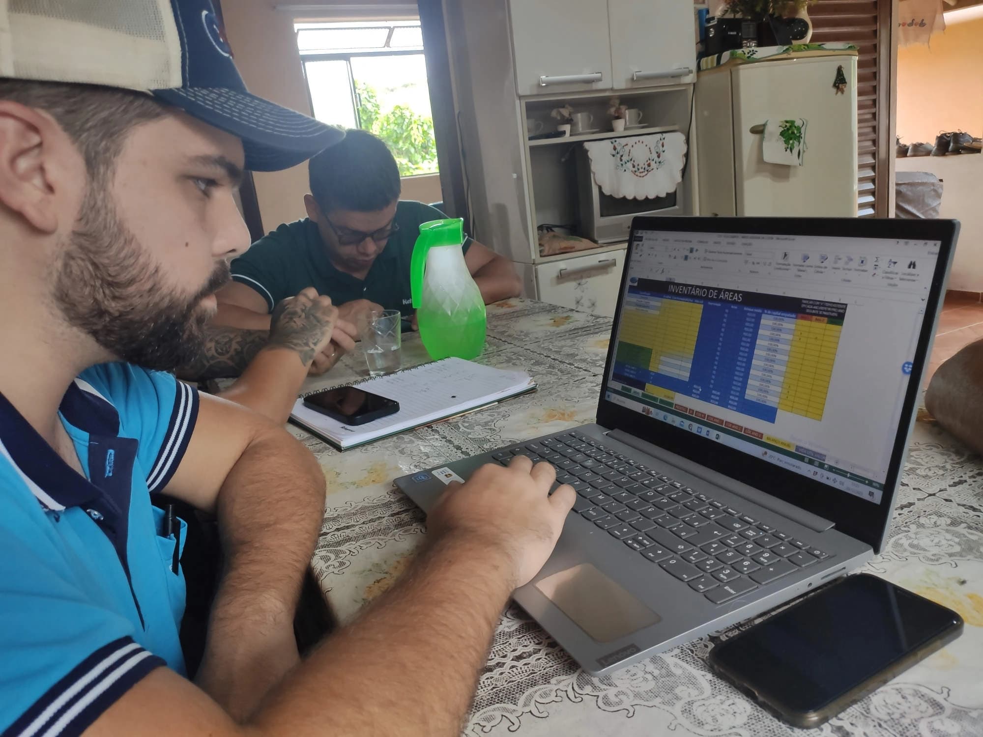 Consultor analisando planilha 'Inventário de Áreas' no notebook, em mesa de cozinha da propriedade rural.
