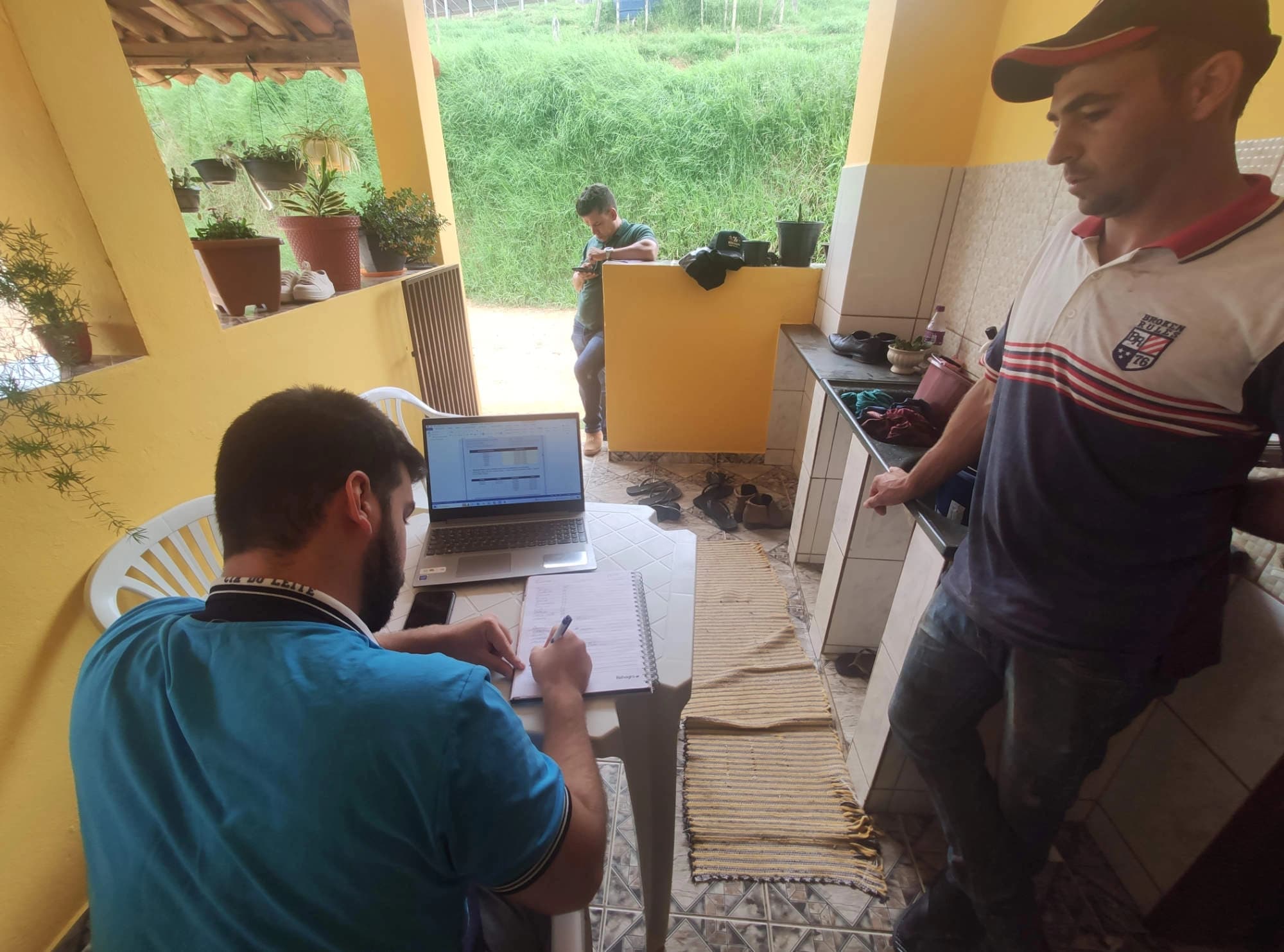 Consultor anotando em caderno ao lado do notebook, durante reunião na varanda da propriedade.